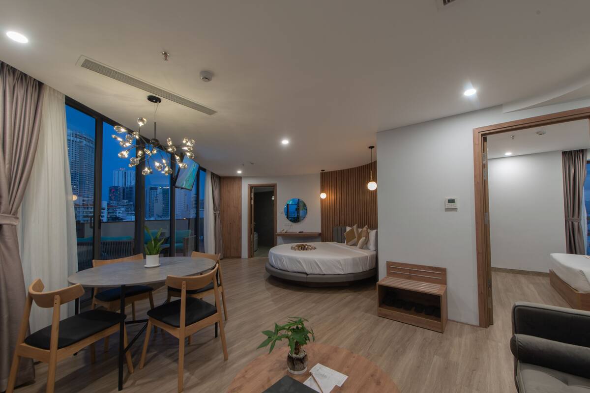 A modern hotel room with a round bed, a dining area with a table and chairs, a small seating area with a sofa, and a view of a cityscape through large windows.