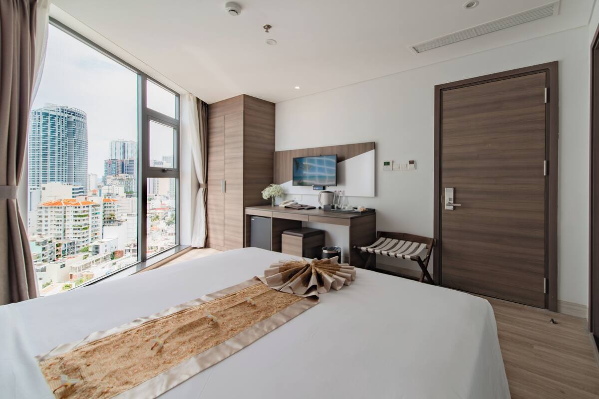 A modern hotel room with a large bed, a desk, a chair, a TV, and a city view.