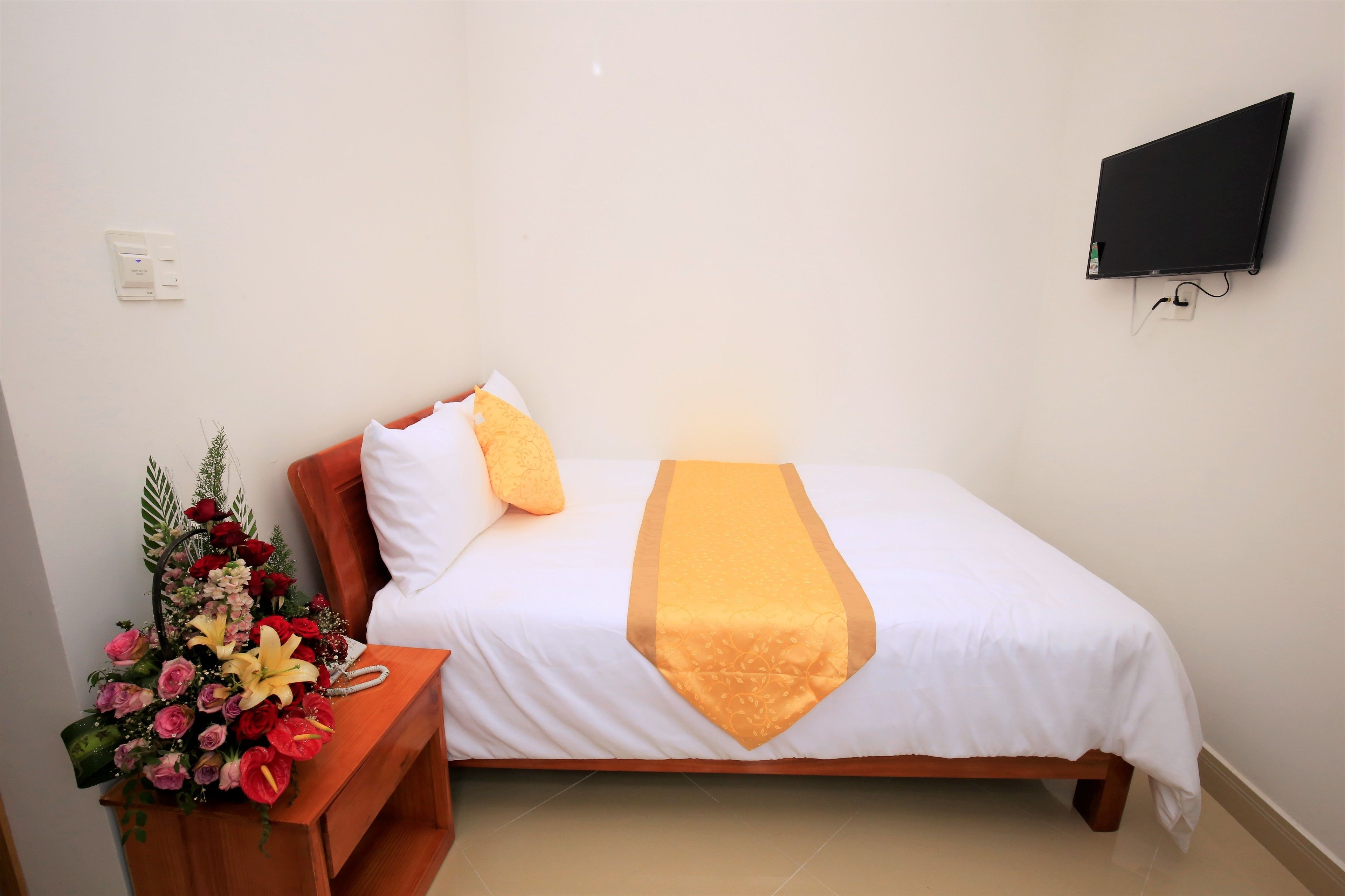 A hotel room with a bed, a bedside table with a floral arrangement, a wall-mounted TV, and a power outlet.