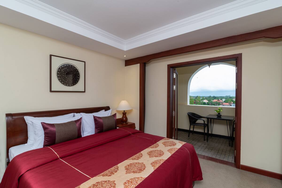 Quarto de hotel com uma cama grande, uma mesa de cabeceira com abajur, um quadro na parede e uma varanda com vista.