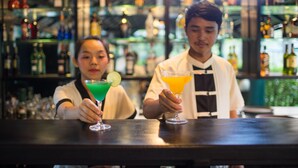 A bar with two people holding drinks, shelves with bottles, and a well-lit interior. - Hotel de la Renaissance (Siem Reap)