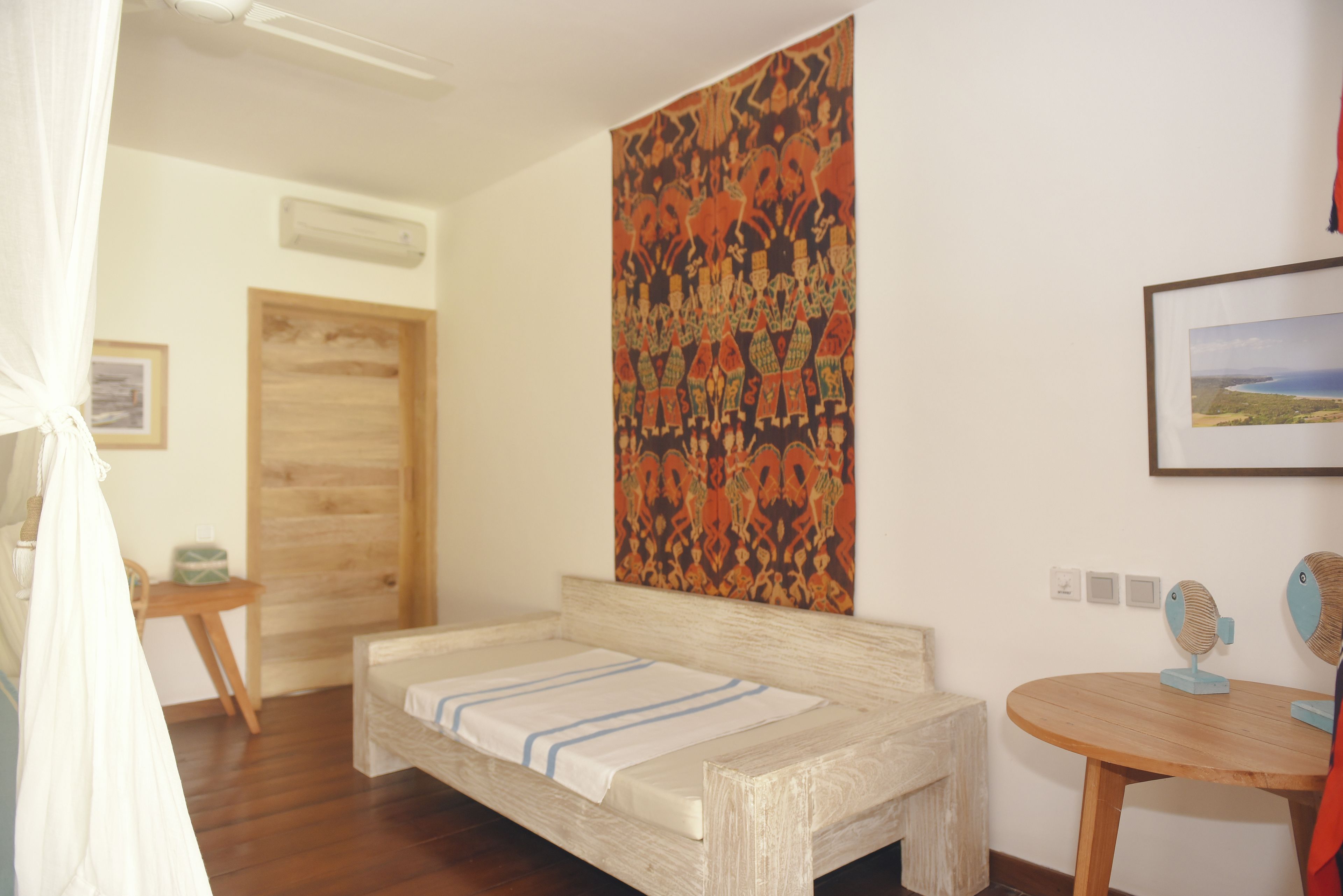 Un dormitorio con una cama de madera, un tapiz, una mesita y una mesa redonda con un ventilador.
