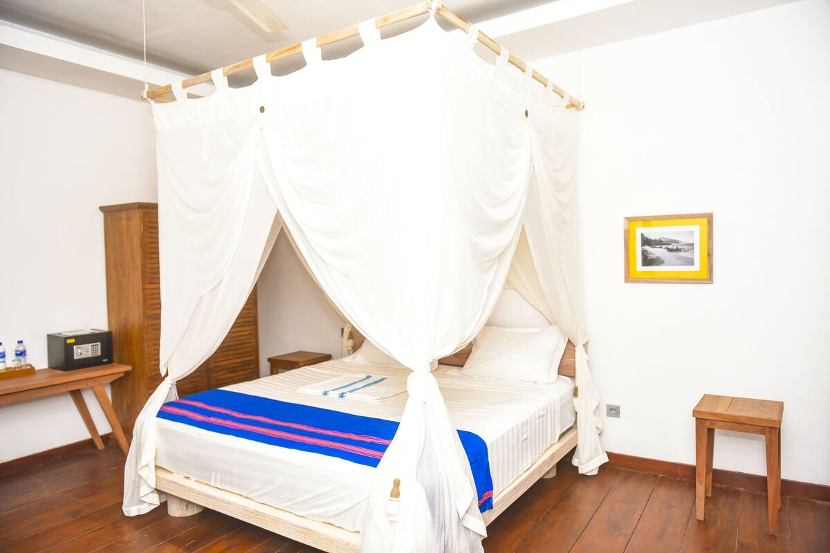 A room with a bed under a white canopy, a wooden nightstand, a small table, and a framed picture on the wall.