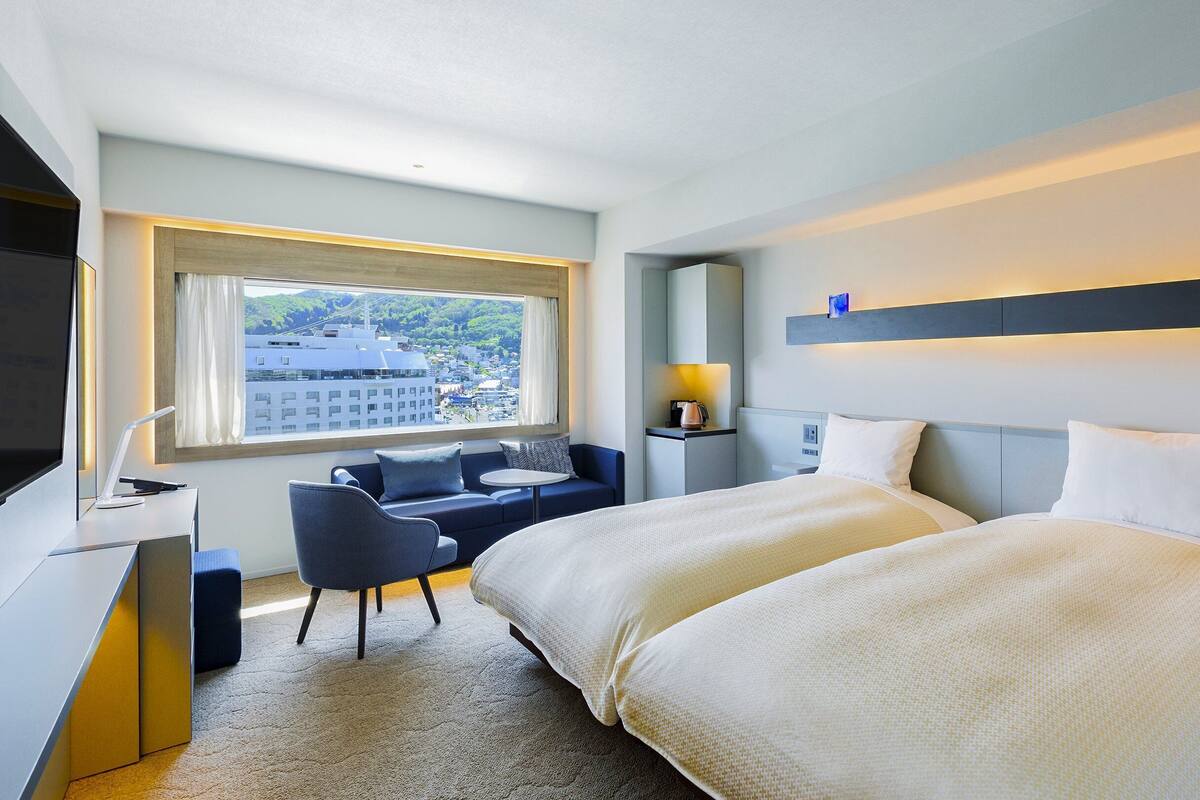 Une chambre d’hôtel avec deux lits, un bureau avec une chaise, une fenêtre donnant sur des immeubles et une table de chevet avec une lampe.