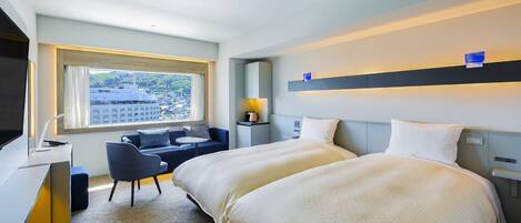 A hotel room with two beds, a desk with a chair, a window with a view of buildings, and a nightstand with a lamp.
