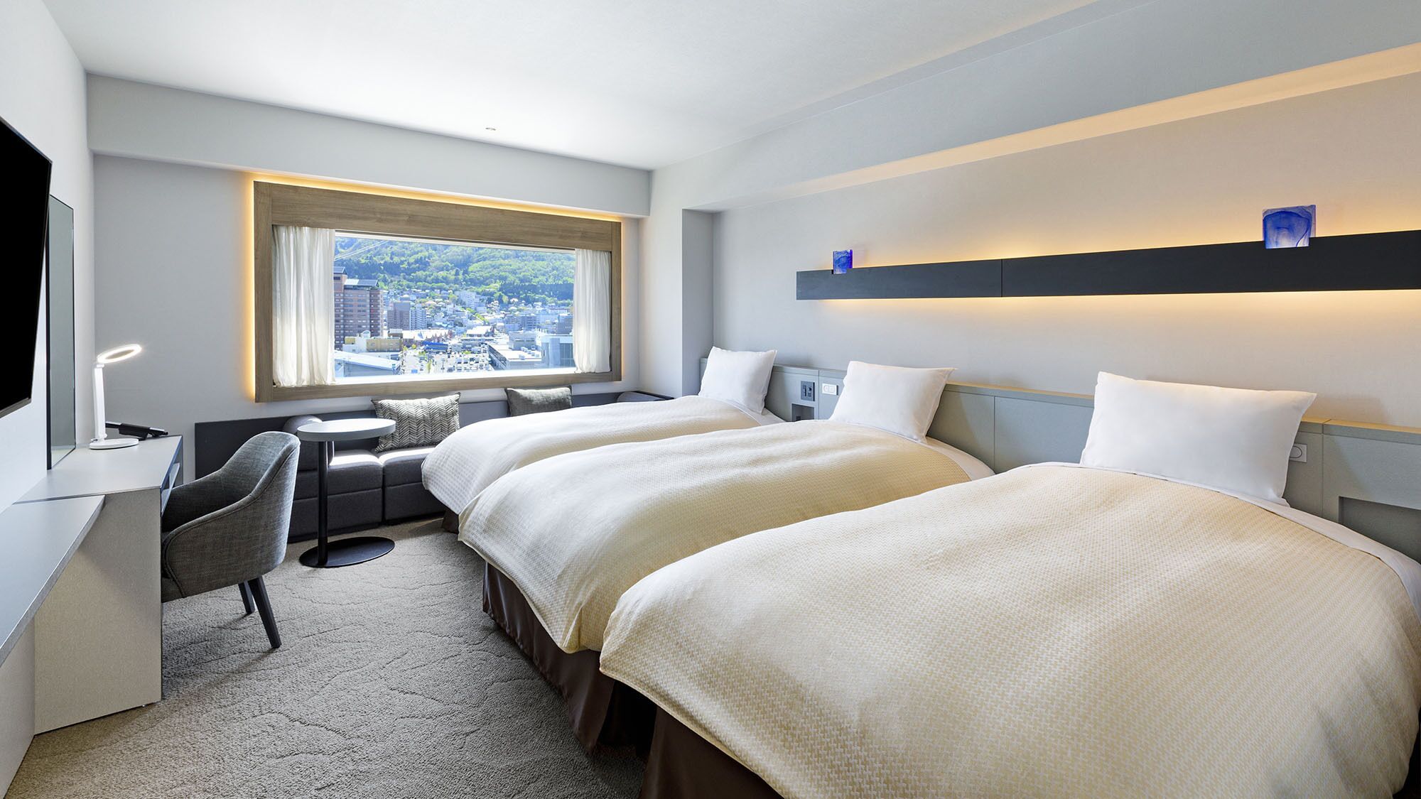 A hotel room with two beds, a desk, a chair, a small table, and a window with a city view.