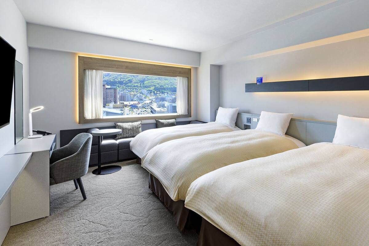 Une chambre d’hôtel avec deux lits, un bureau, une chaise, une petite table et une fenêtre donnant sur la ville.