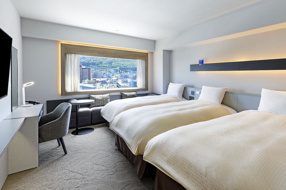 A hotel room with two beds, a desk, a chair, a small table, and a window with a city view.