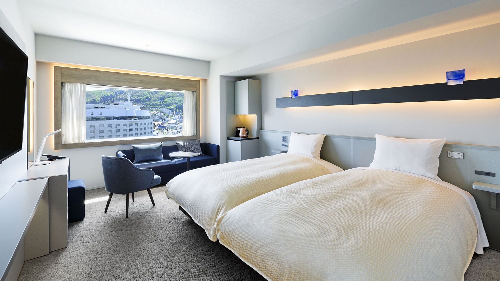 A hotel room with a bed, a desk, a chair, a window with a view of buildings, and a nightstand with a lamp.