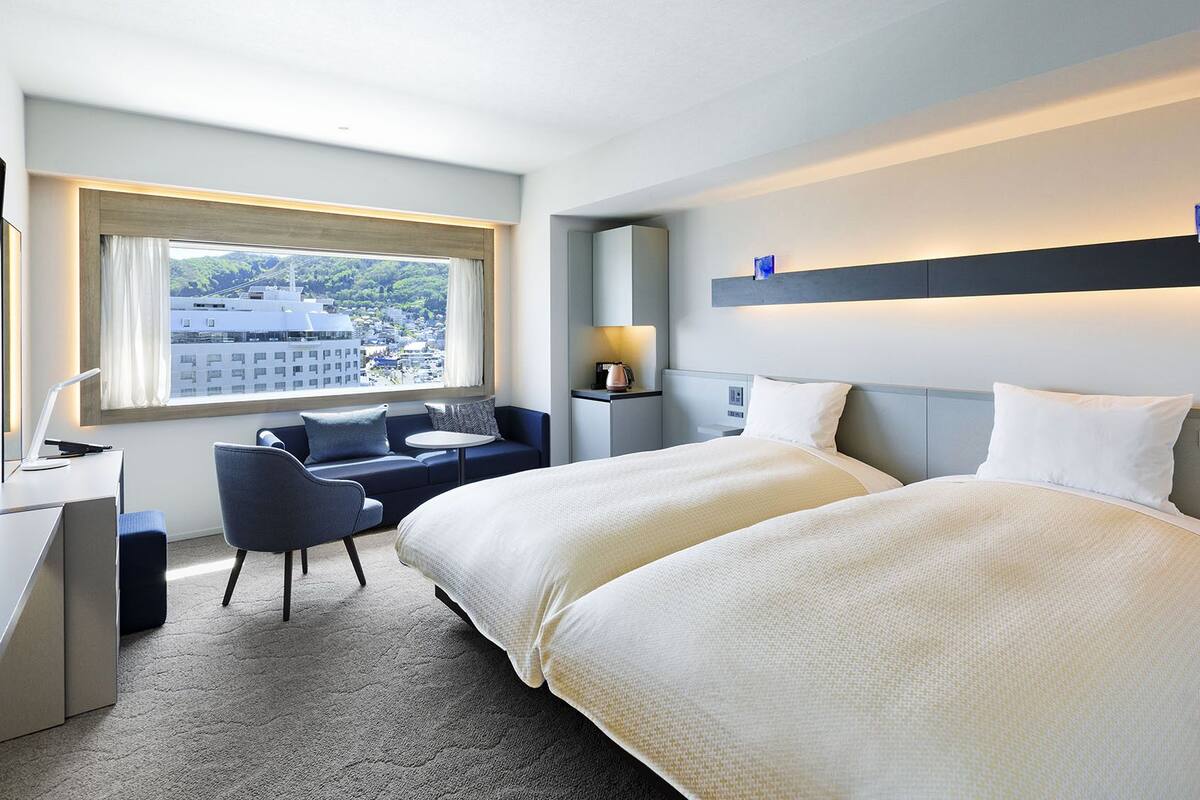 Une chambre d’hôtel comprenant un lit, un bureau, une chaise, une fenêtre donnant sur des immeubles et une table de chevet avec une lampe.