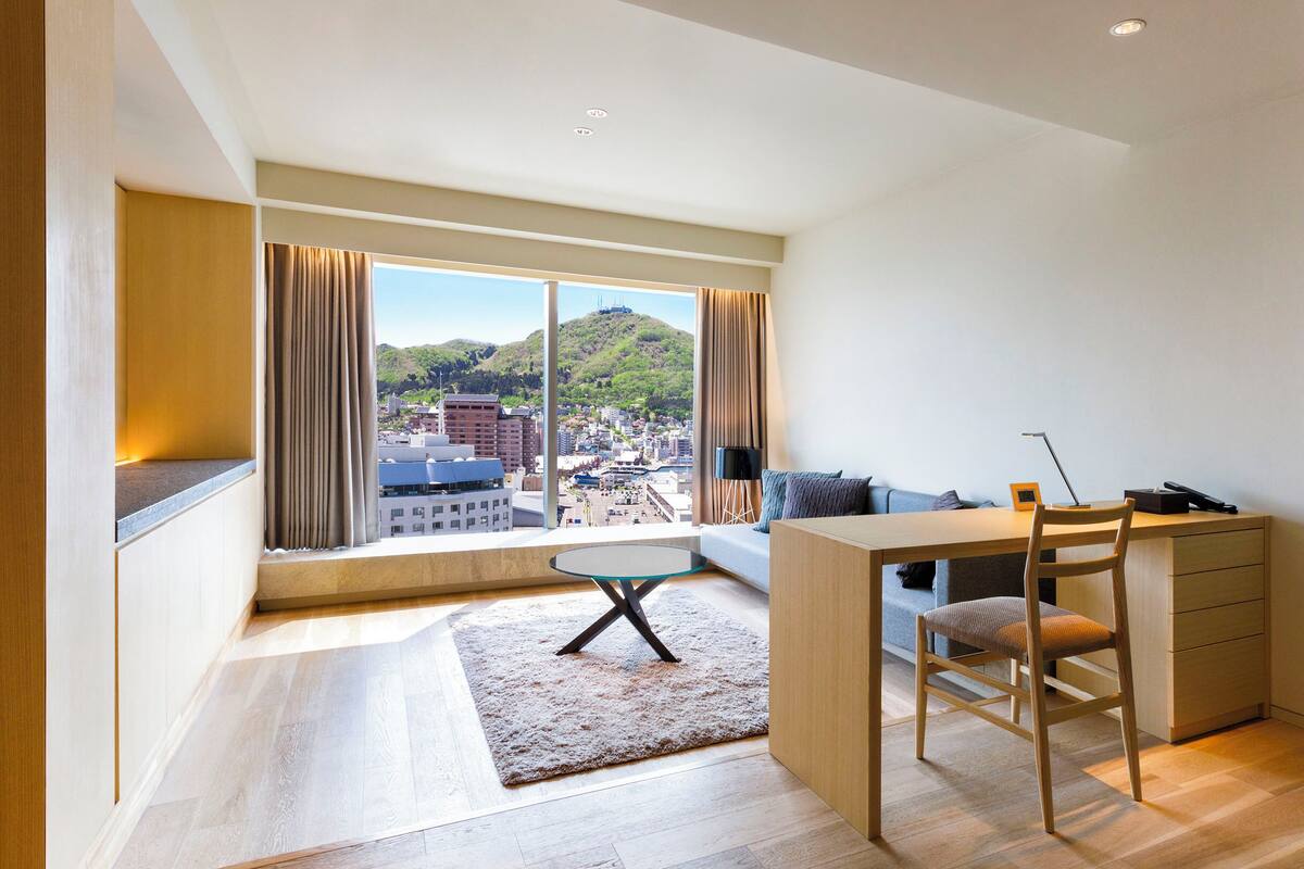 Une chambre d’hôtel moderne dotée d’une grande fenêtre donnant sur la ville, d’un bureau avec une chaise et d’un canapé.