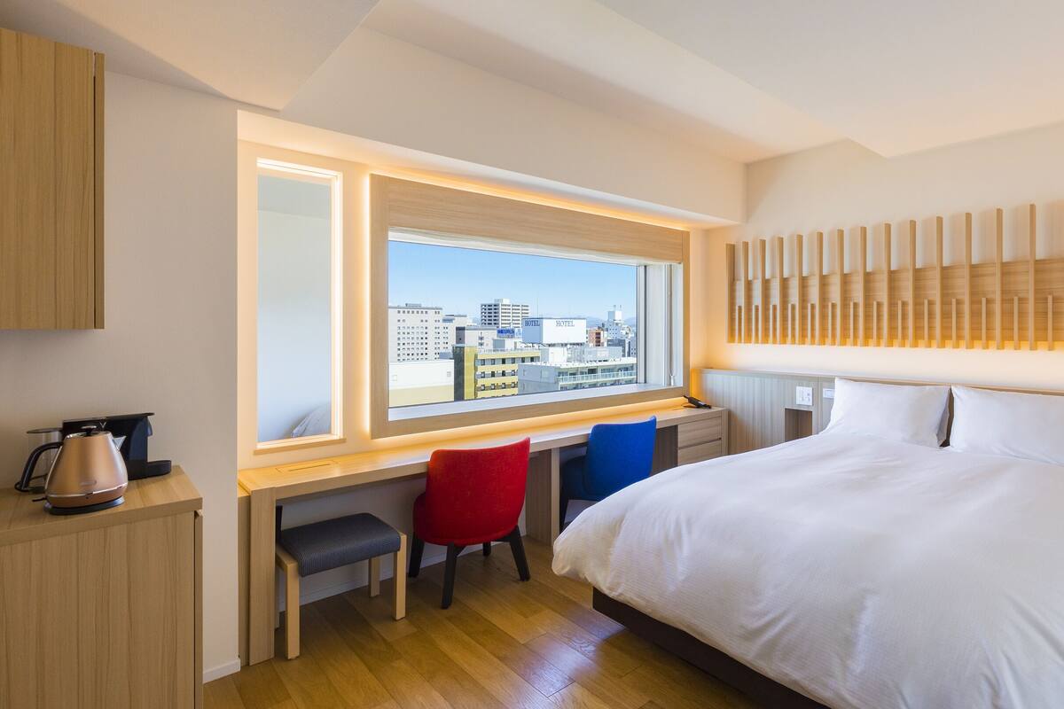 Une chambre d’hôtel avec un grand lit, un bureau avec une chaise, une fenêtre donnant sur la ville et un luminaire moderne au-dessus du lit.