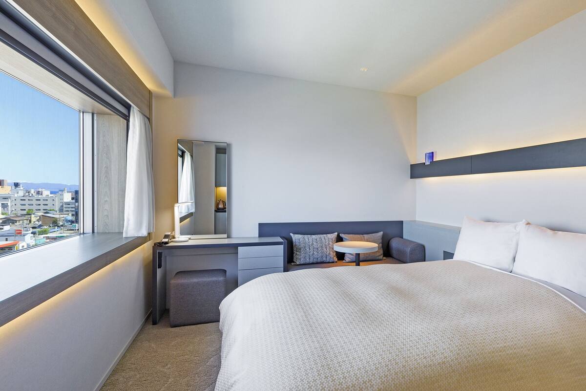 Une chambre d’hôtel moderne, dotée d’un grand lit, d’un bureau, d’une chaise et d’une petite table ronde. Une fenêtre offre une vue sur la ville.