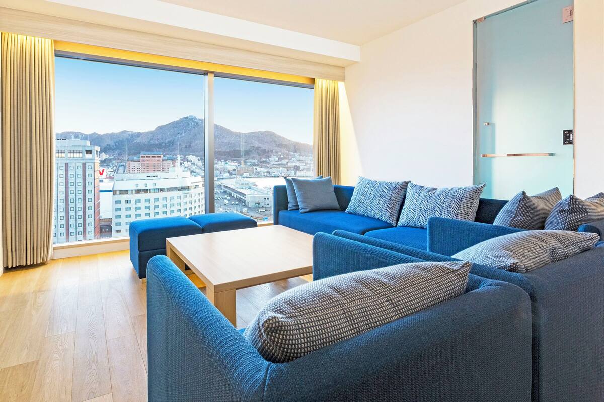 Une chambre d’hôtel moderne dotée d’une grande fenêtre donnant sur la ville, d’un canapé bleu, d’une table basse en bois et d’un téléviseur à écran plat fixé au mur.