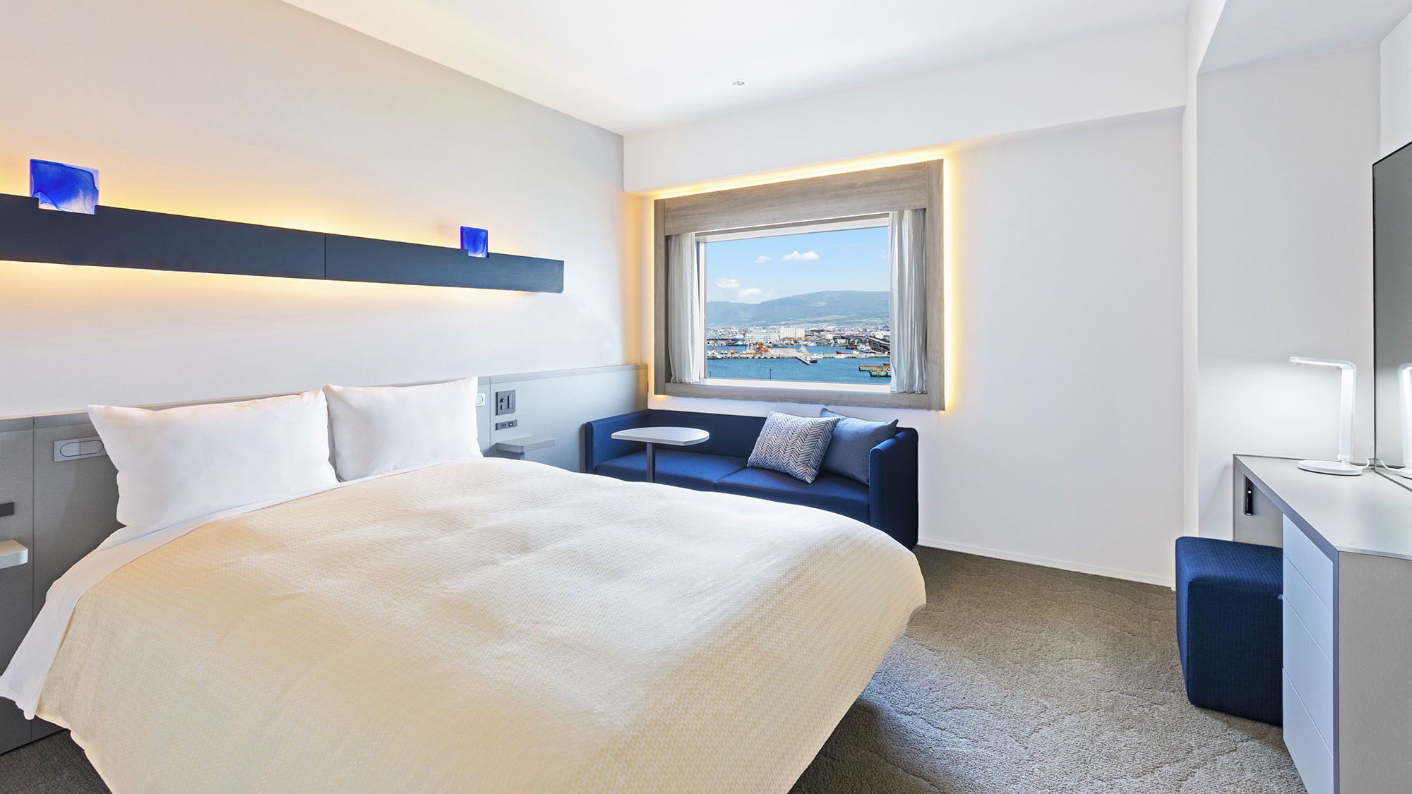 A hotel room with a bed, a seating area, a desk, and a window with a view of a coastal city.