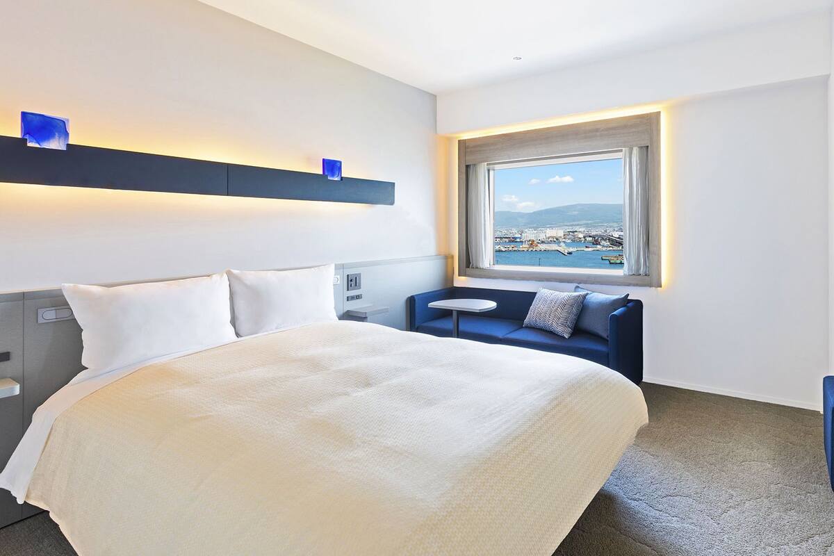 Une chambre d’hôtel comprenant un lit, un coin salon, un bureau et une fenêtre donnant sur une ville côtière.