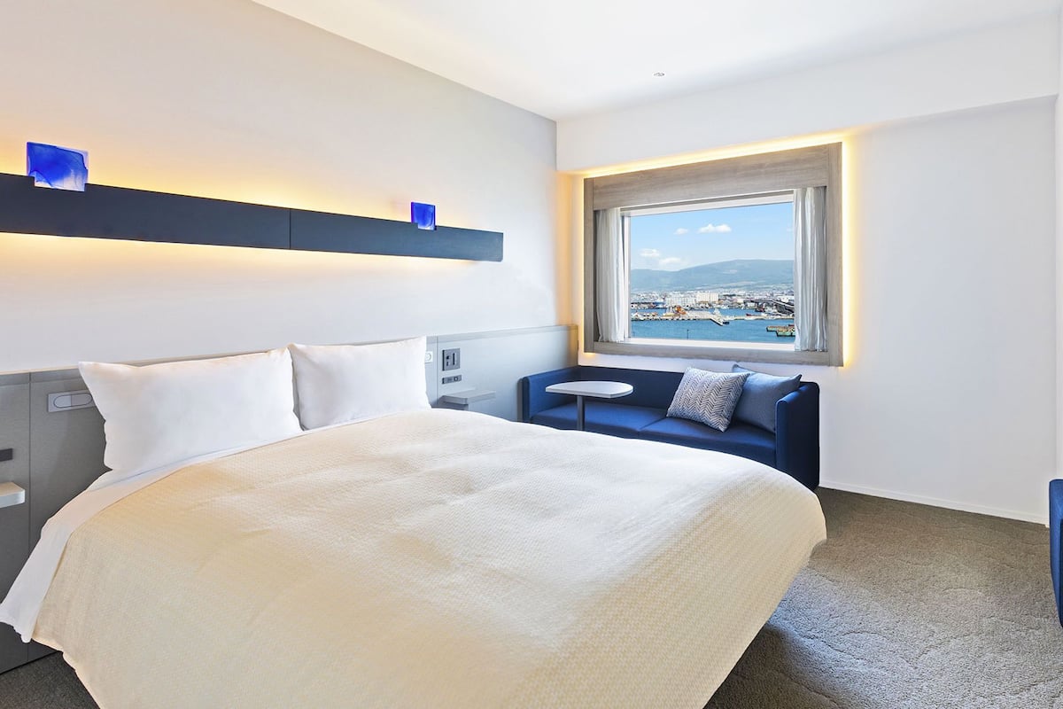 A hotel room with a bed, a seating area, a desk, and a window with a view of a coastal city.