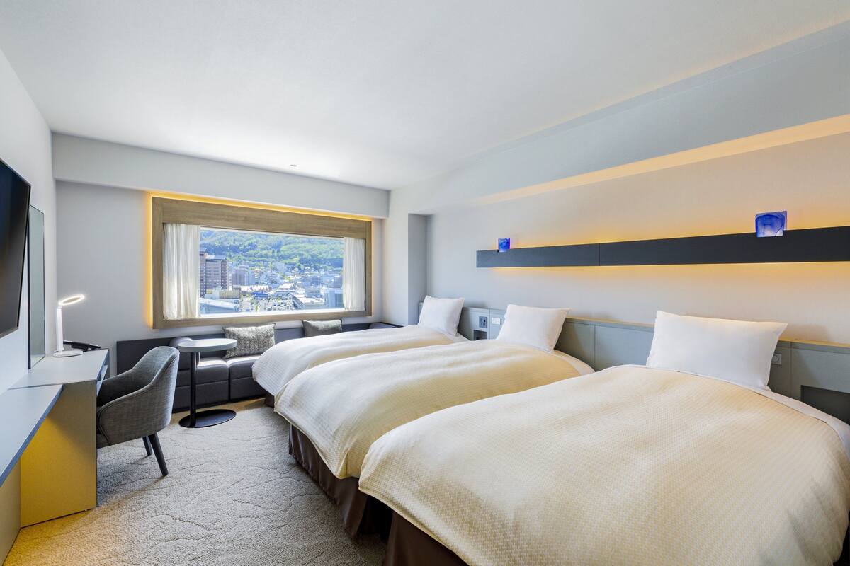 Une chambre d’hôtel avec deux lits, un bureau, une chaise, une petite table et une fenêtre donnant sur la ville.