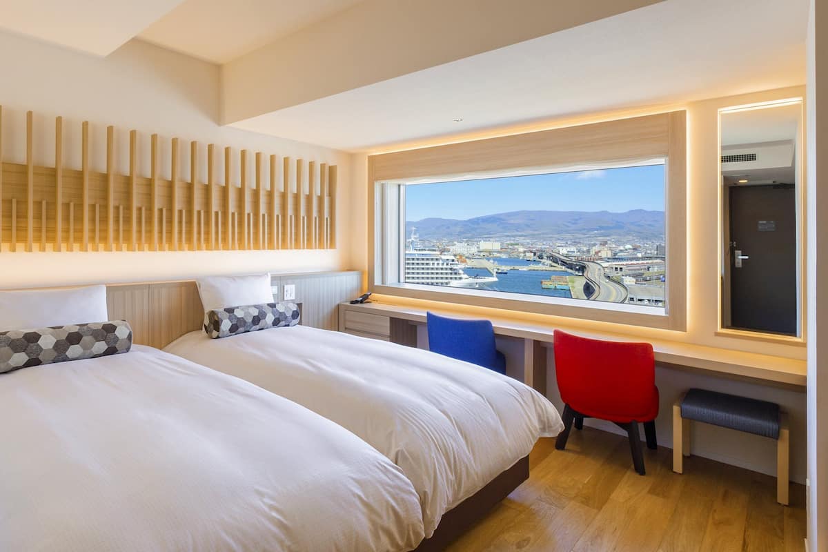 Une chambre d’hôtel moderne avec deux lits, un bureau et une grande fenêtre donnant sur la ville.
