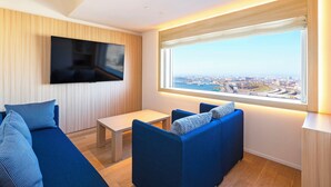 Ein modernes Wohnzimmer mit großem Fernseher, blauen Sofas, einem kleinen Tisch und Blick auf die Stadt durch ein großes Fenster.