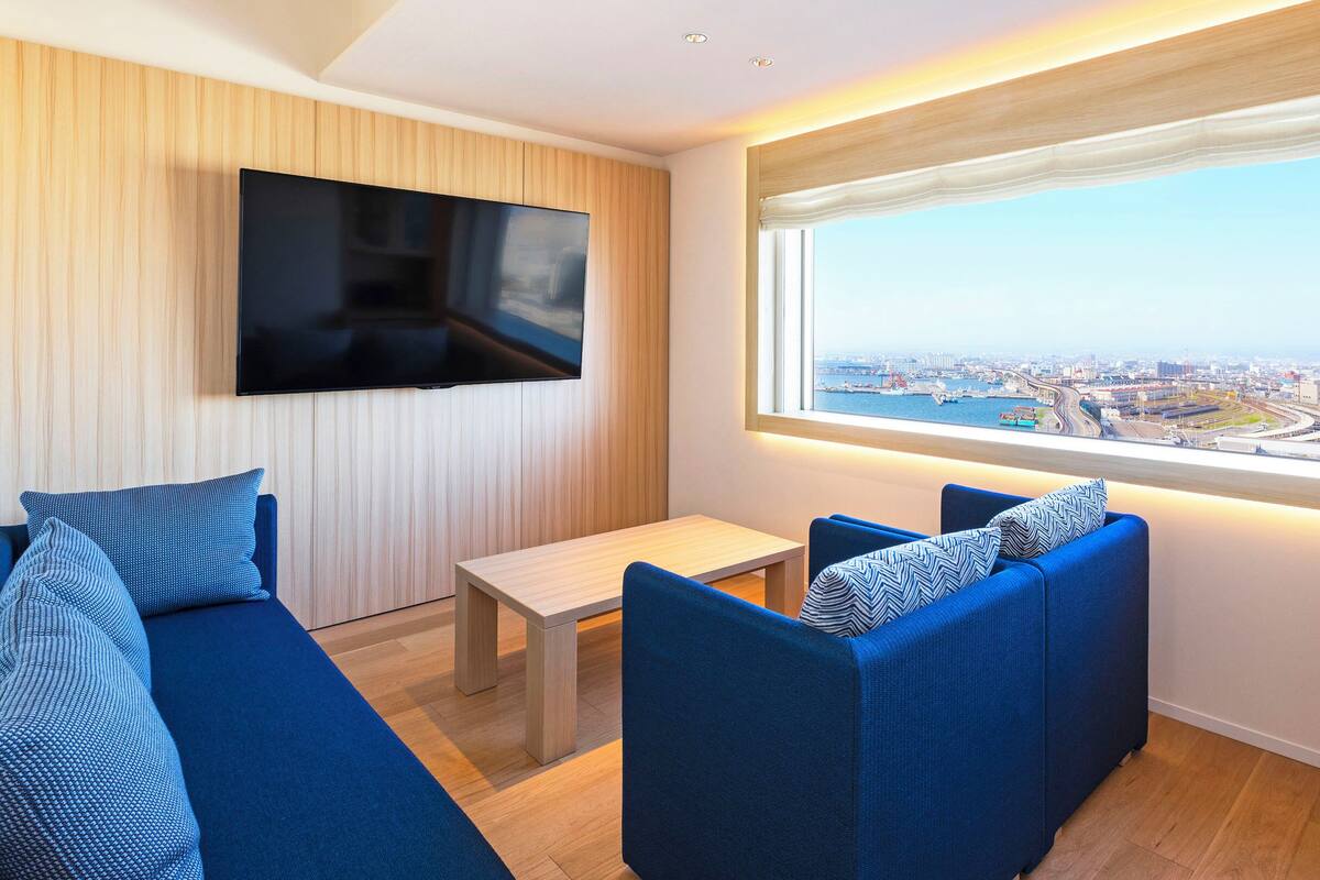 Un salon moderne avec une grande télévision, des canapés bleus, une petite table et une vue sur la ville par une grande fenêtre.