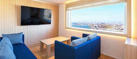 A modern living room with a large TV, blue sofas, a small table, and a city view through a large window.