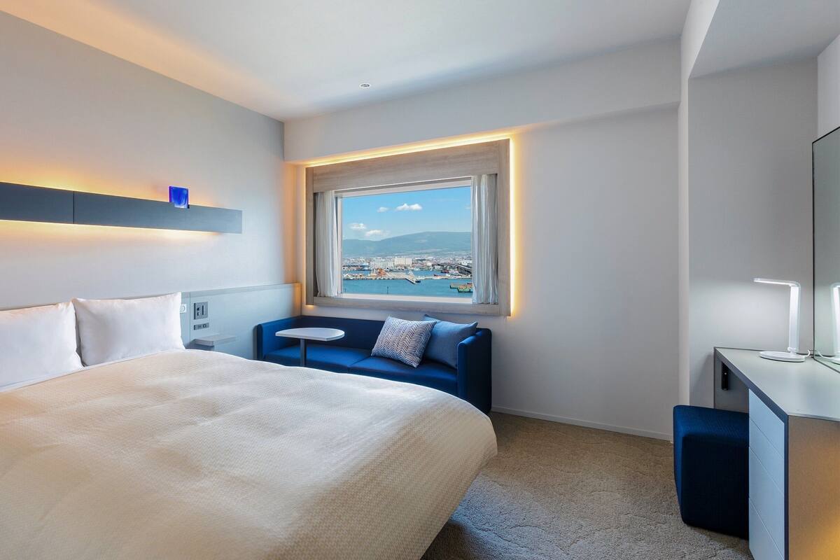 Une chambre d’hôtel moderne avec un grand lit, une petite table, un canapé et une vue sur le paysage urbain par la fenêtre.