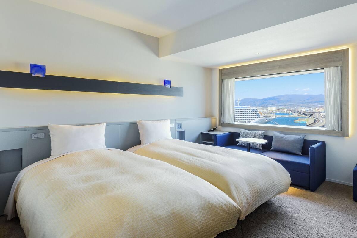 Une chambre d’hôtel moderne dotée d’un grand lit, d’un coin salon avec vue sur la ville et d’une fenêtre donnant sur le port.