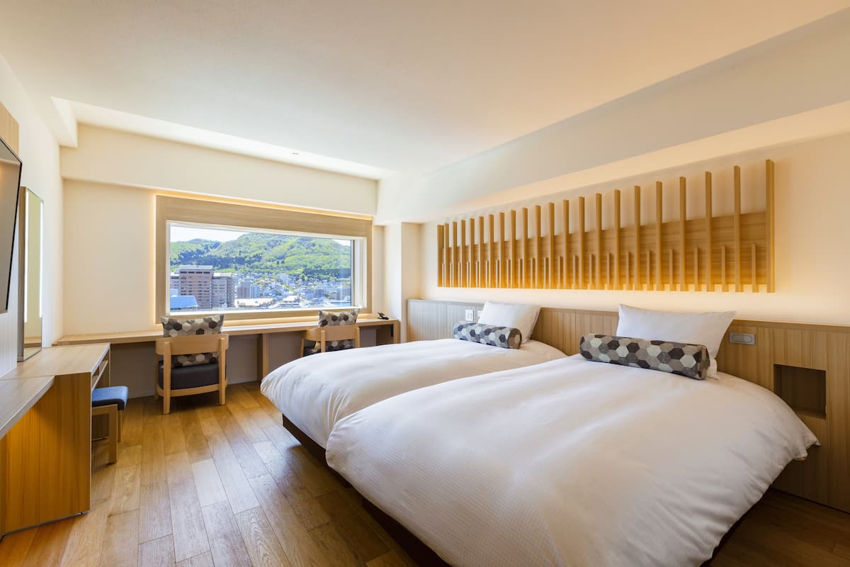 Une chambre d’hôtel moderne avec deux lits, un bureau et une grande fenêtre donnant sur la ville.