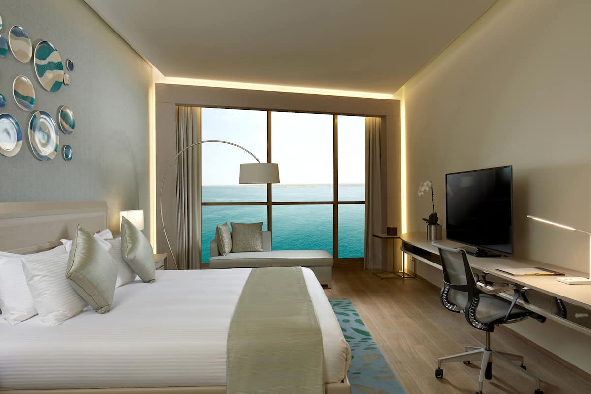 A hotel room with a bed, a desk, a television, and a large window with a view of the sea.