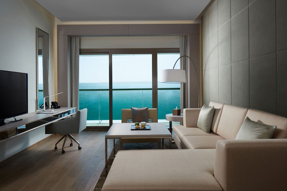 A modern hotel room with a large window, a flat-screen TV, a sofa, a coffee table, and a desk with a lamp.