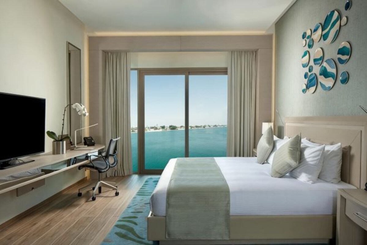 A hotel room with a bed, a desk, a television, and a view of the sea.