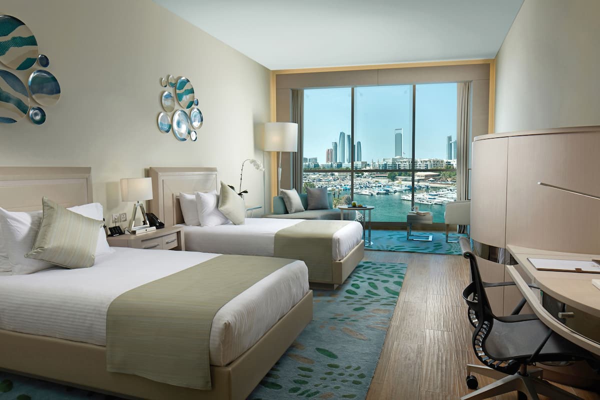 A hotel room with two beds, a desk, and a view of a marina and city skyline.