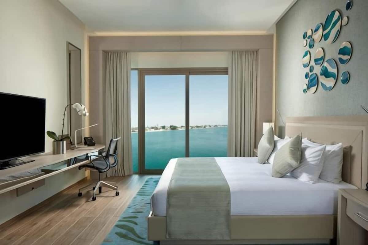 A hotel room with a bed, a desk, a television, and a view of the sea.