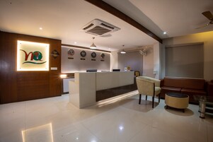 Zona de recepción de hotel con diseño moderno, que incluye un mostrador de recepción, un área de estar con sofá y sillones, y una pared con relojes y un logotipo.