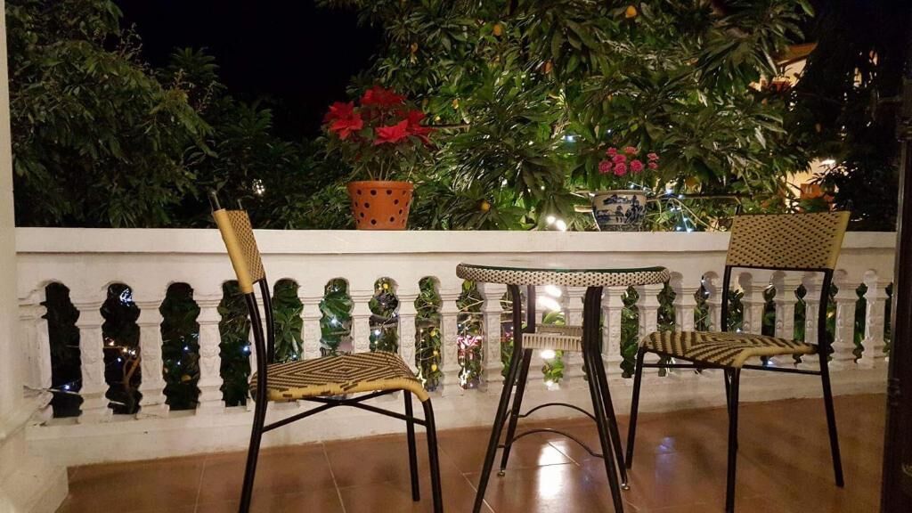 A balcony with two wicker chairs and a small table, surrounded by a white balustrade and lush greenery.