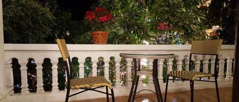 A balcony with two wicker chairs and a small table, surrounded by a white balustrade and lush greenery.