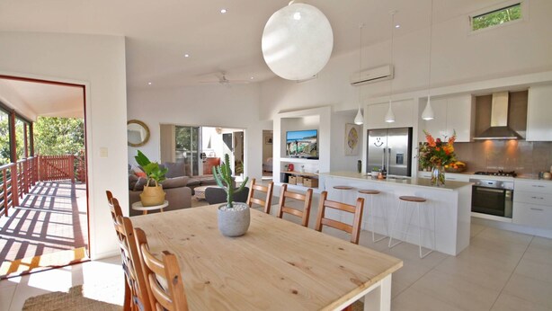 A modern kitchen with white cabinets, a large island, and a dining area with wooden chairs and a table. - Stunning Home With Spectacular Views (Coolum Beach)