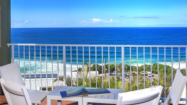 A balcony with white chairs and a table, overlooking a beach and the ocean.