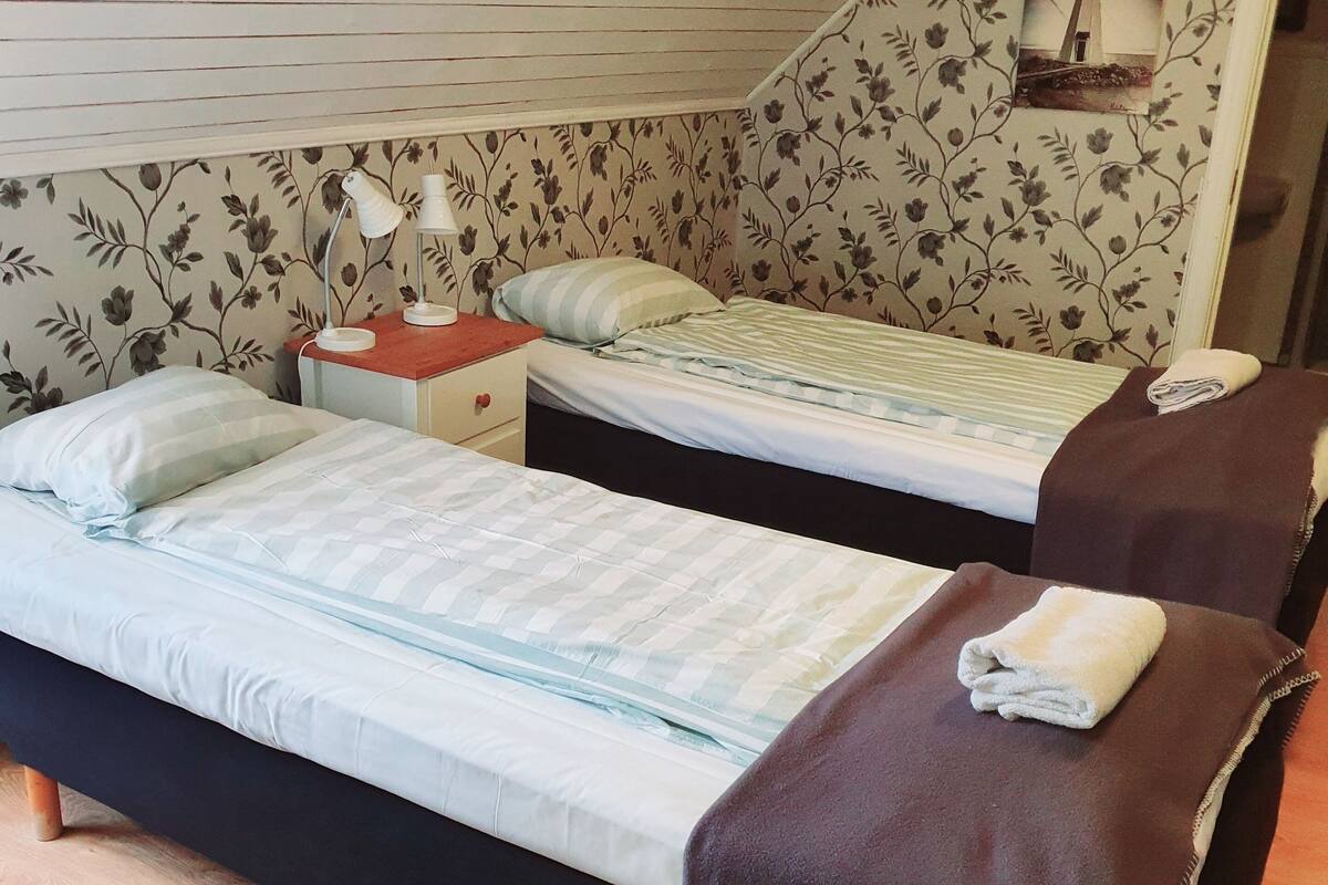 Un dormitorio con dos camas, suelo de madera, papel tapiz floral, una mesita de noche con lámpara y un cuadro enmarcado en la pared.