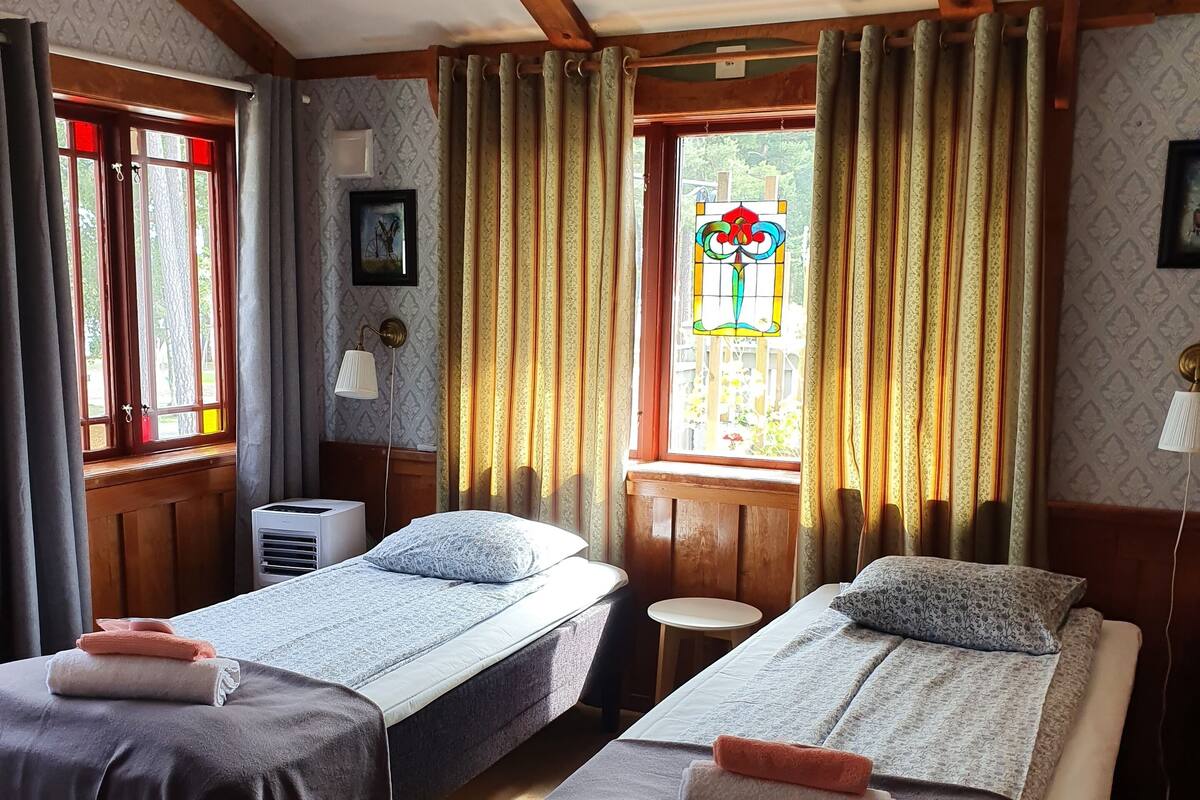 Una habitación con dos camas, una mesita, una lámpara y un ventanal con cortinas.