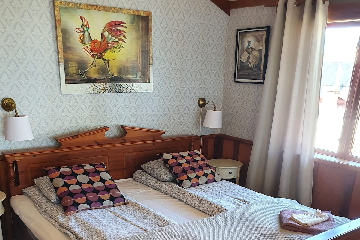 Una cama de madera con cabecera, dos mesitas de noche y un tocador. Hay dos lámparas sobre el tocador, un cuadro enmarcado en la pared y una ventana con cortinas.