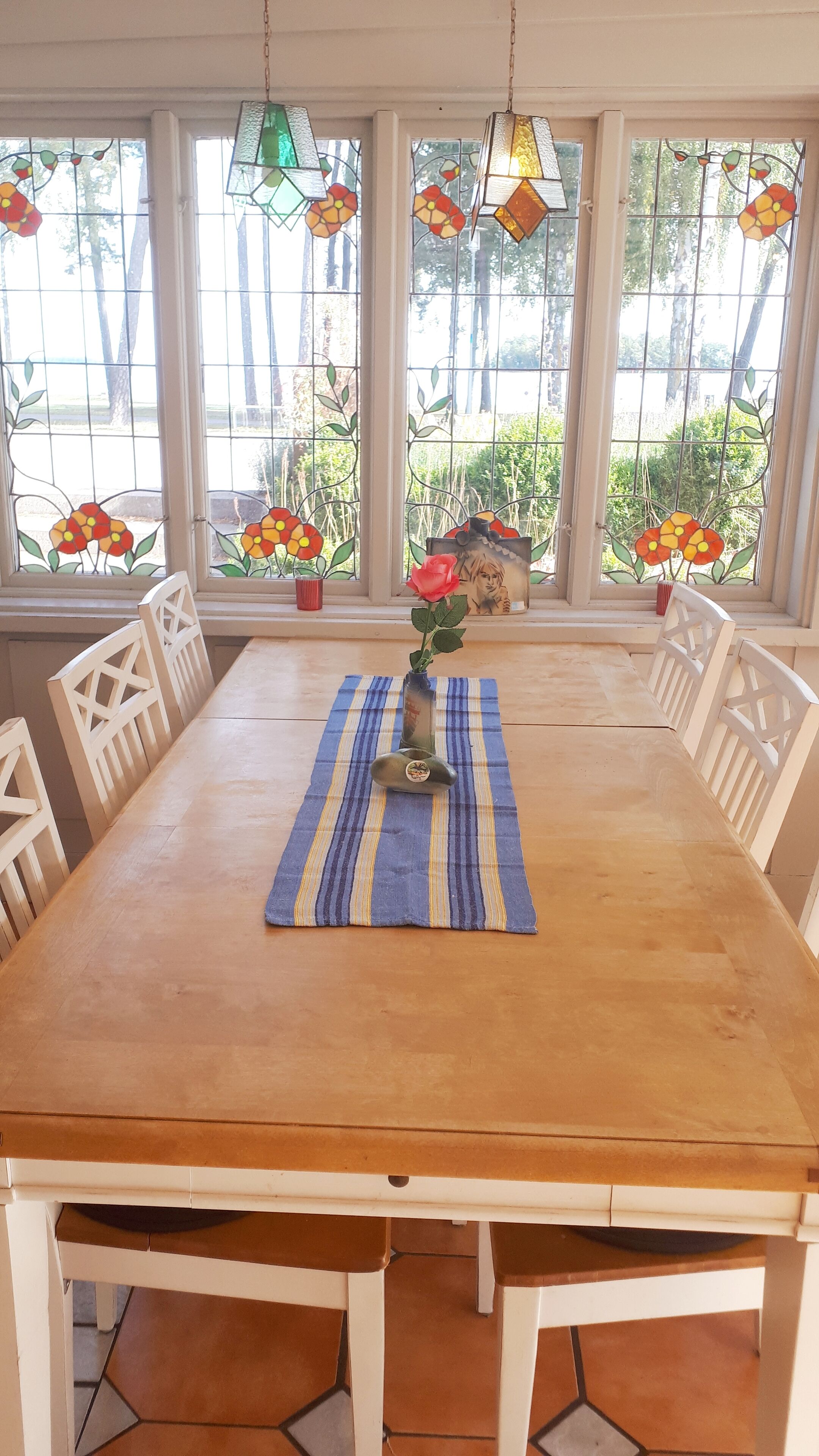 Un coin repas avec une table en bois, des chaises blanches, un set de table bleu à rayures et un vase contenant une seule rose rouge.
