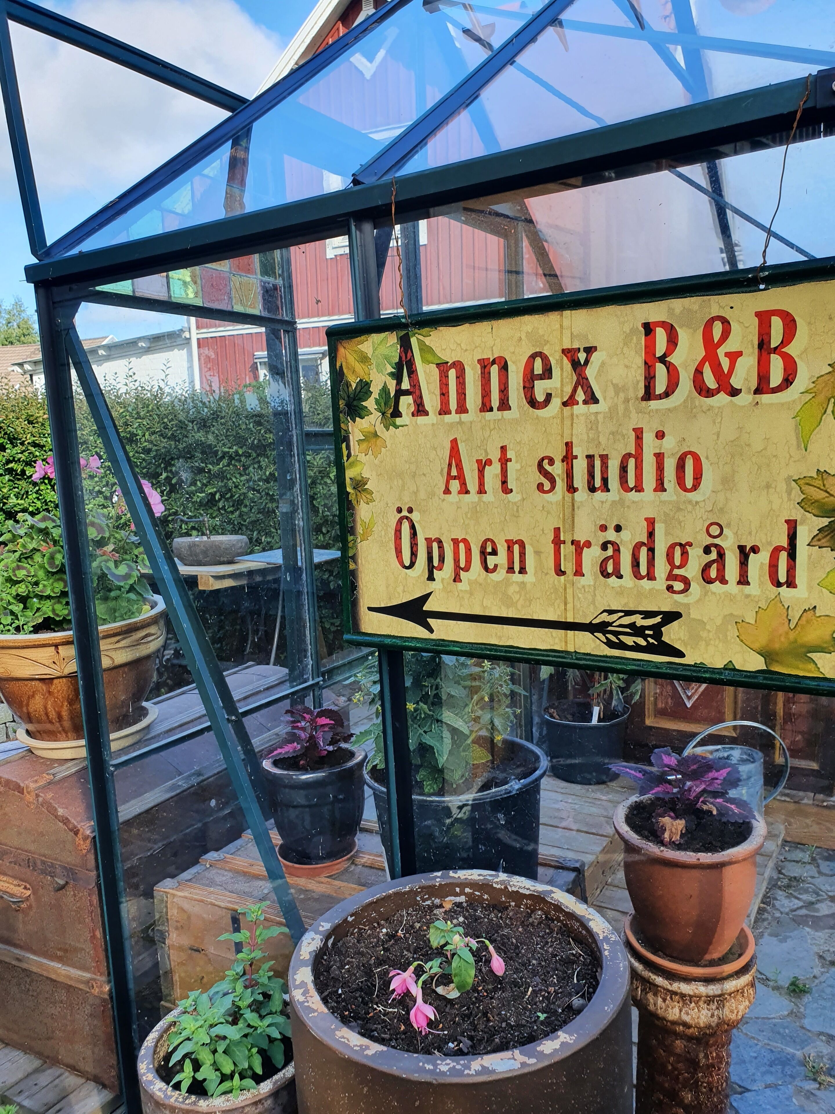 Un hivernacle amb un cartell que diu "Annex B&B Art studio Öppen trädgård" i plantes en testos a l'interior.