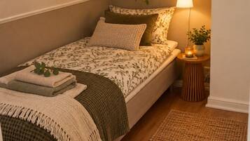 Una cama individual con una colcha estampada, una lámpara de noche, una mesita con una planta y una alfombra tejida sobre un suelo de madera.