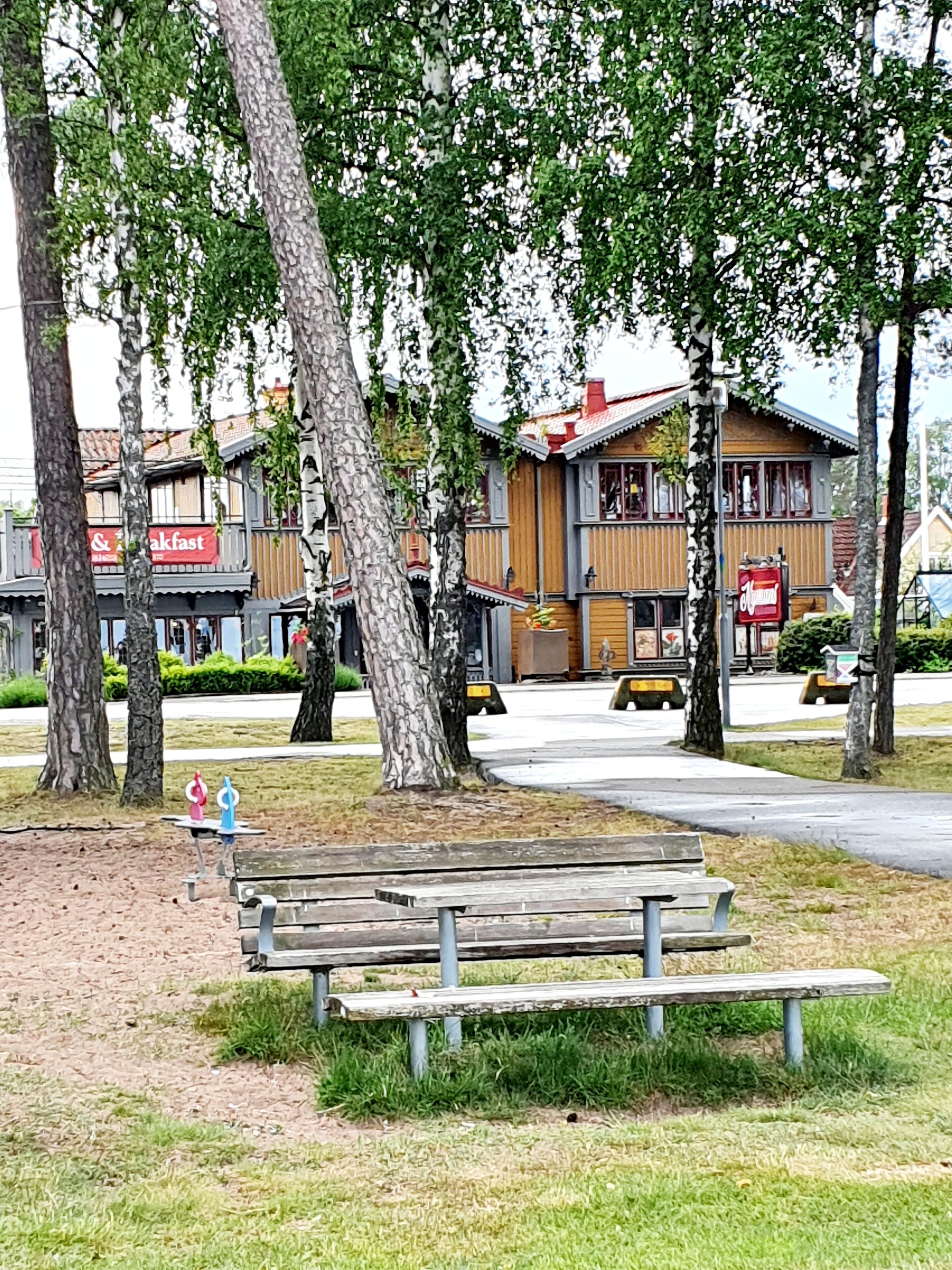 Un parque con un banco de madera, árboles y un edificio con toldo rojo.