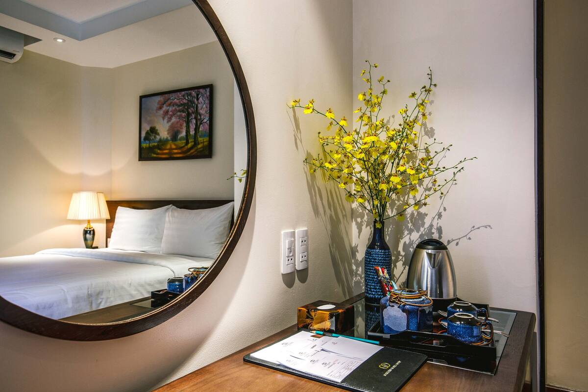 A hotel room with a bed, a desk with a lamp, a vase with flowers, a coffee maker, and a mirror reflecting the room.