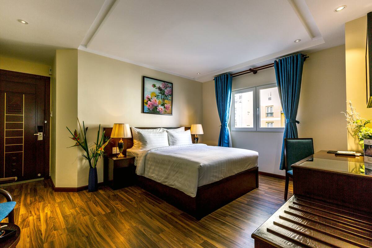 A hotel room with a wooden floor, a bed with white linens, a desk with a chair, a window with blue curtains, and a painting on the wall.