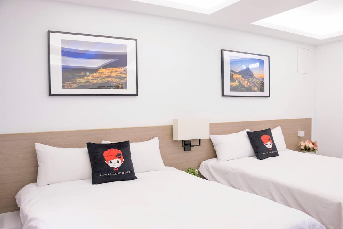 Habitación de hotel con dos camas, cada una con un cojín que presenta un personaje de dibujos animados. En la pared hay cuadros enmarcados, una lámpara de noche y un pequeño jarrón con flores.