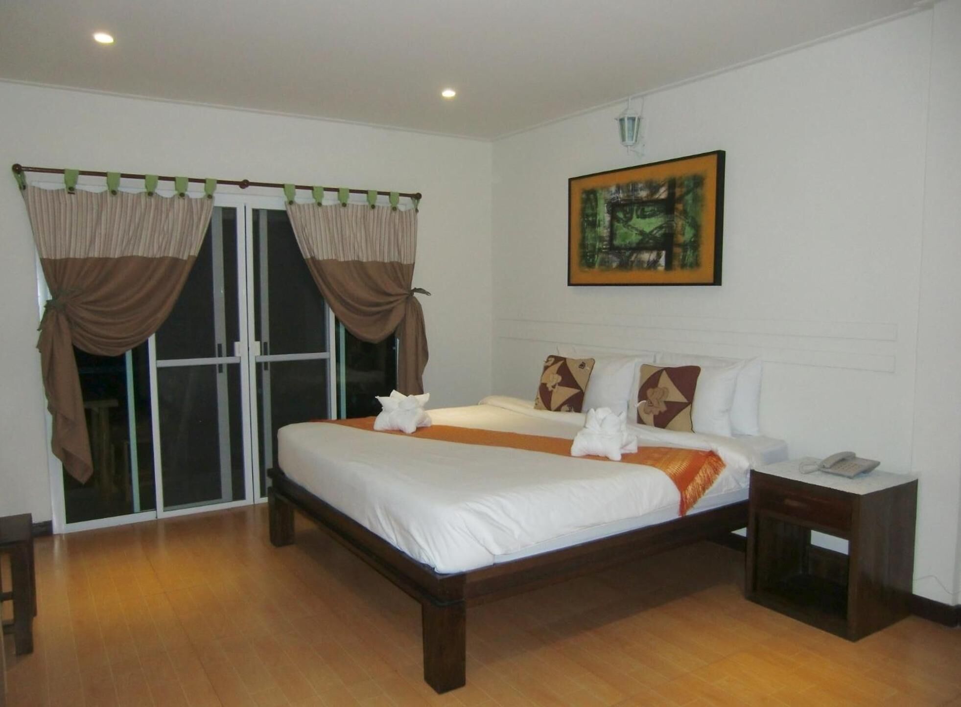 A hotel room with a large bed, two bedside tables, a nightstand, a painting on the wall, and a sliding glass door with curtains.